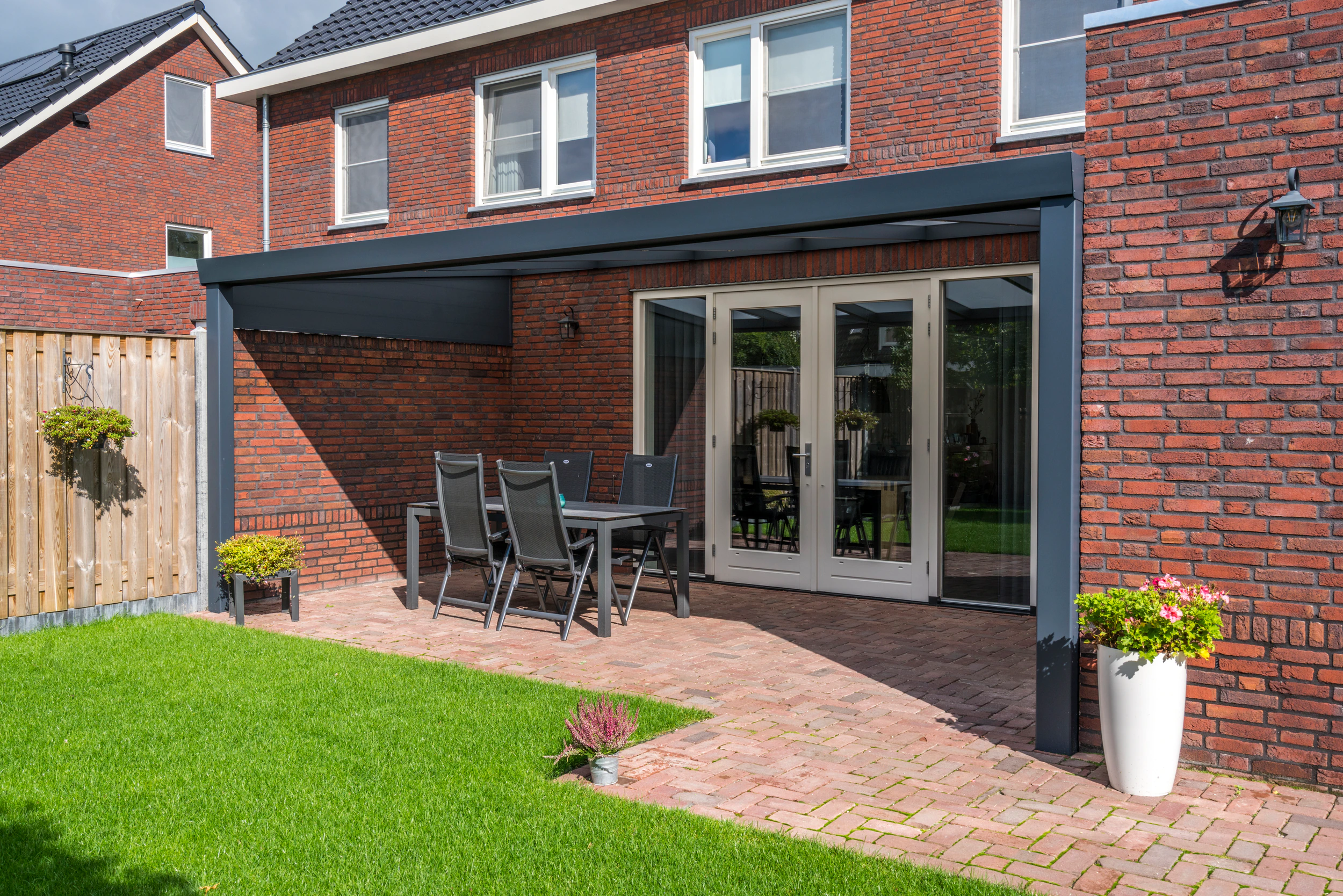 Aluminium side wall installed on a veranda behind a red-brick Dutch home with outdoor dining chairs and manicured lawn