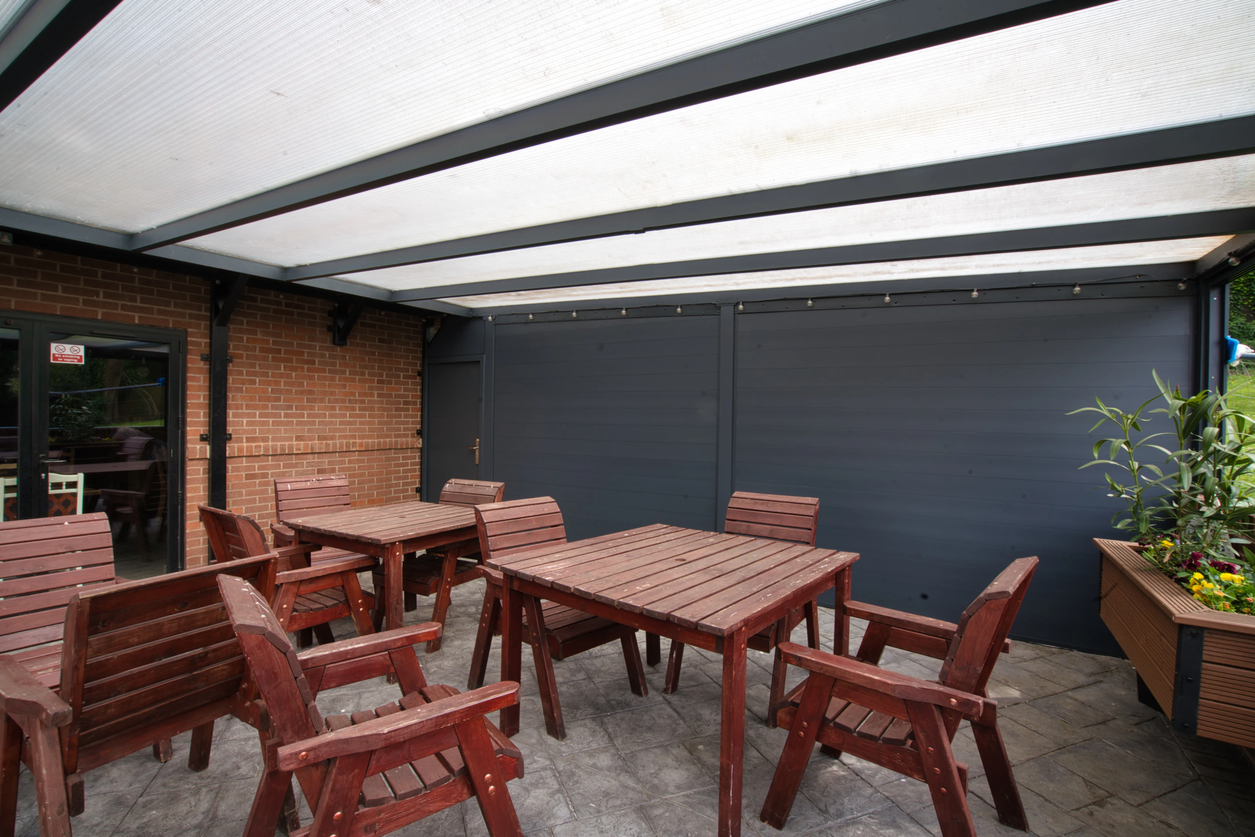 Aluminium side wall in anthracite grey shielding a covered outdoor seating area with wooden pub-style tables and chairs