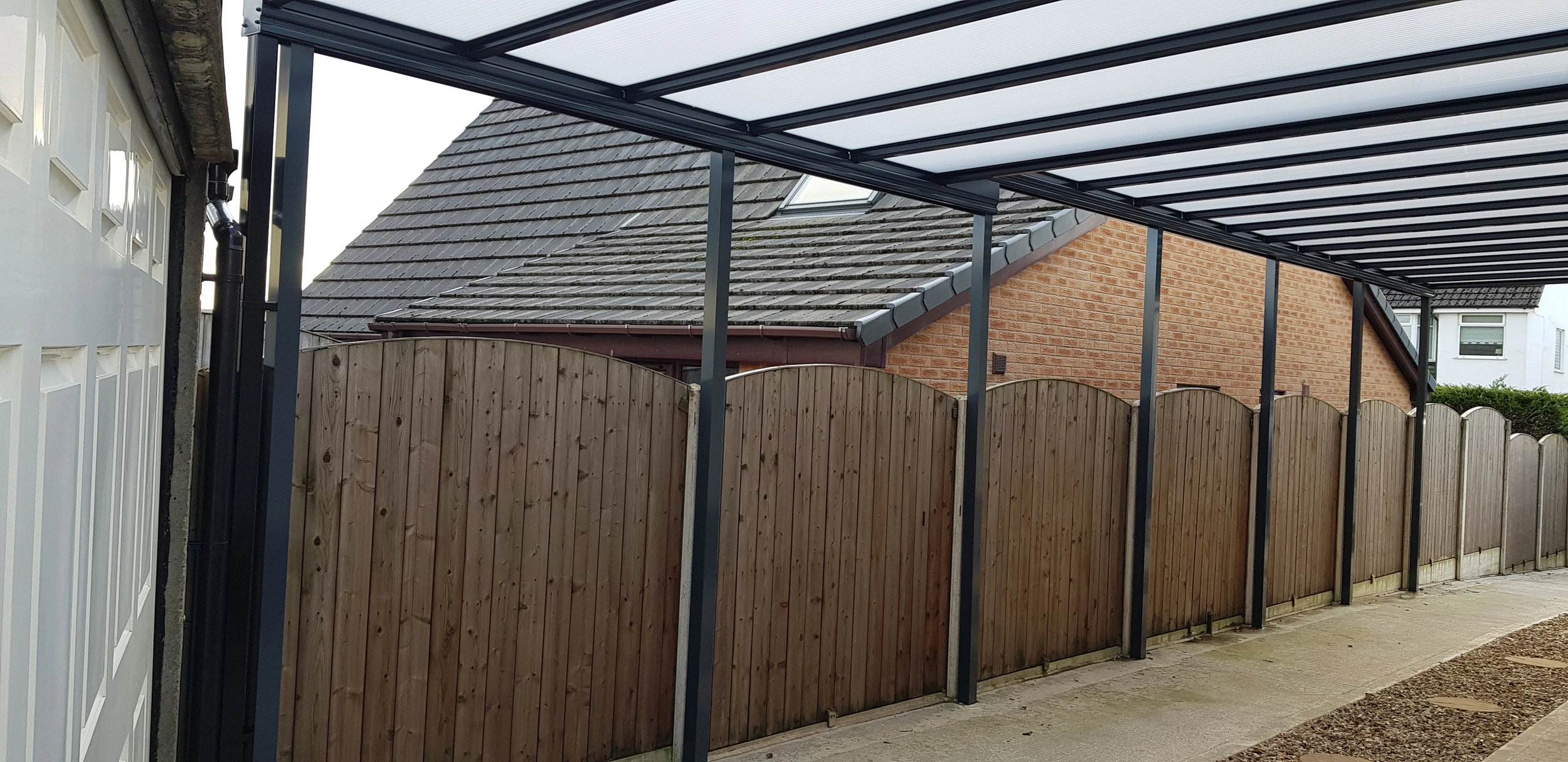 Harbour anthracite carport extended along a long driveway beside arched wooden fencing