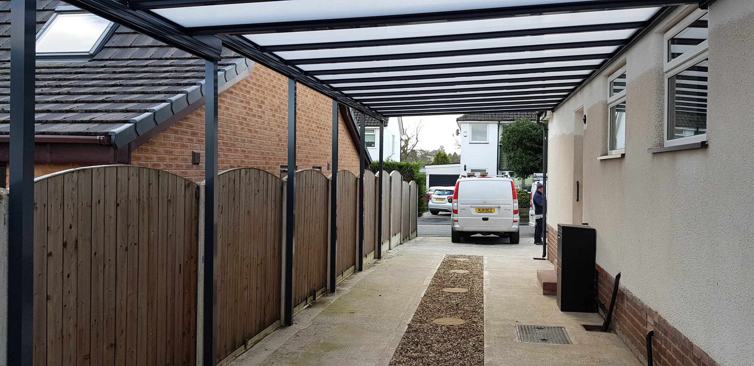 Harbour carport covering a driveway with white van and arched panel fencing