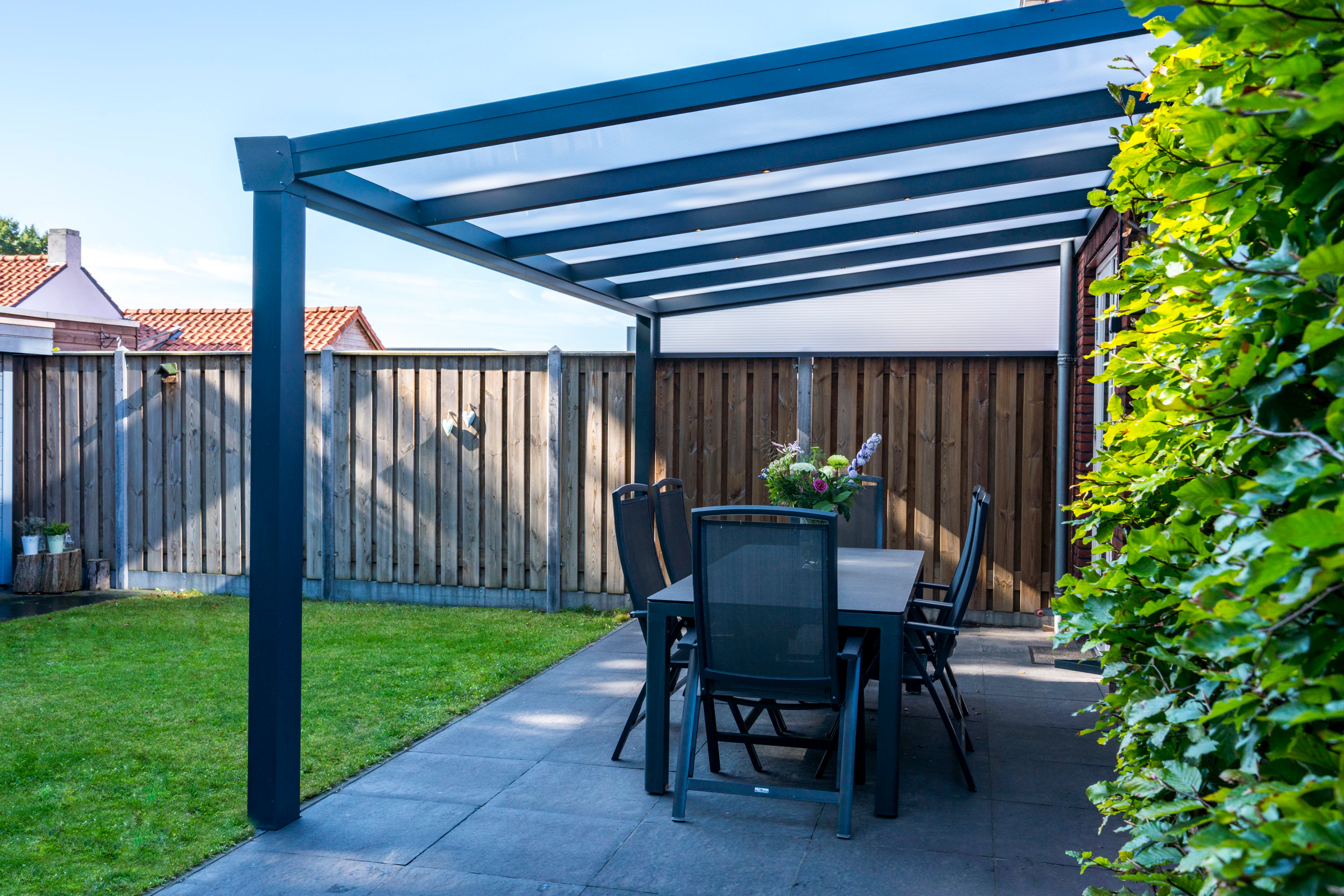 Anthracite aluminium pergola with polycarbonate side wall creating a sheltered outdoor dining area beside a wooden fence