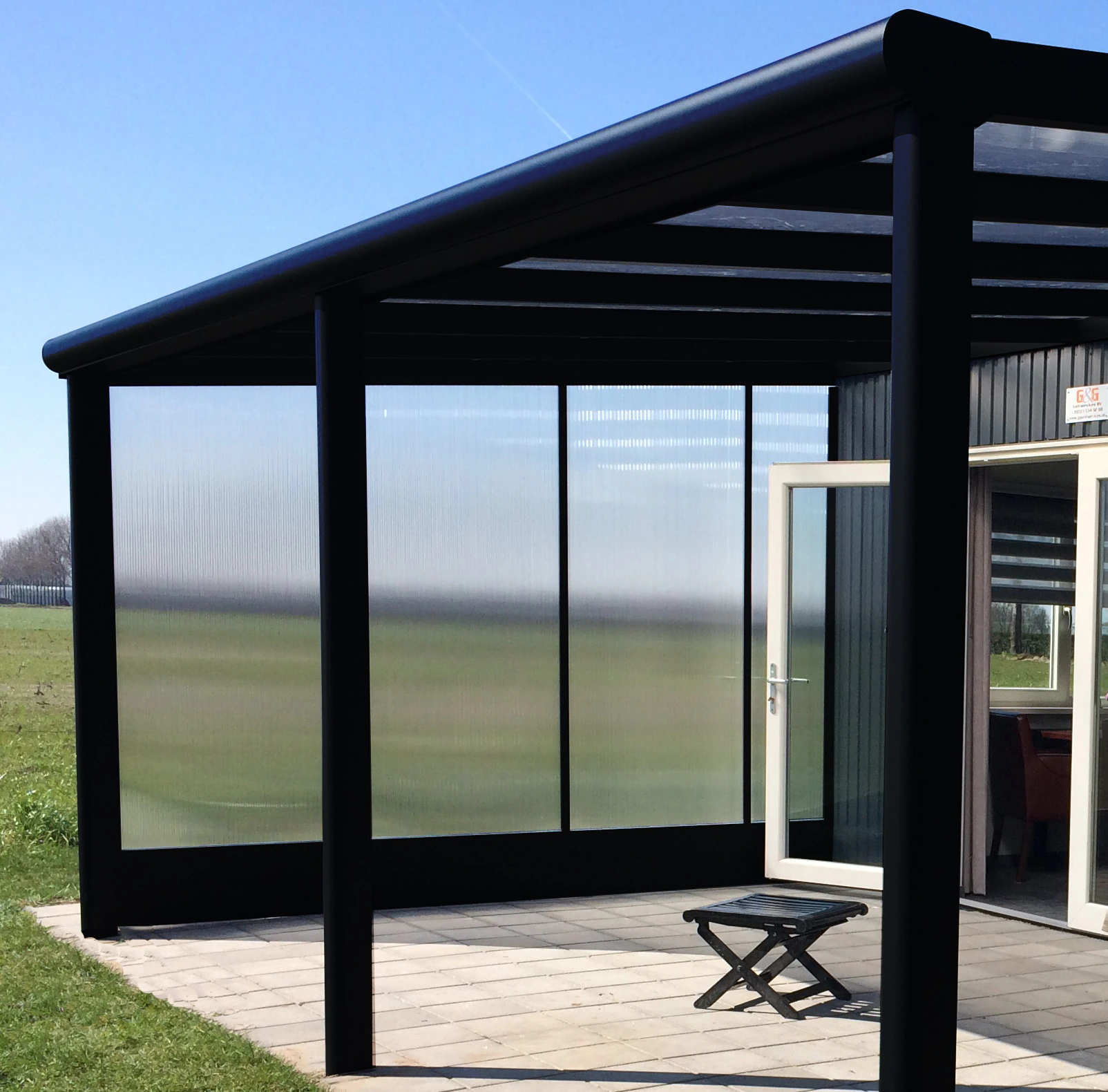 Black-framed polycarbonate side wall enclosing a veranda with countryside field view, paved patio, and folding stool