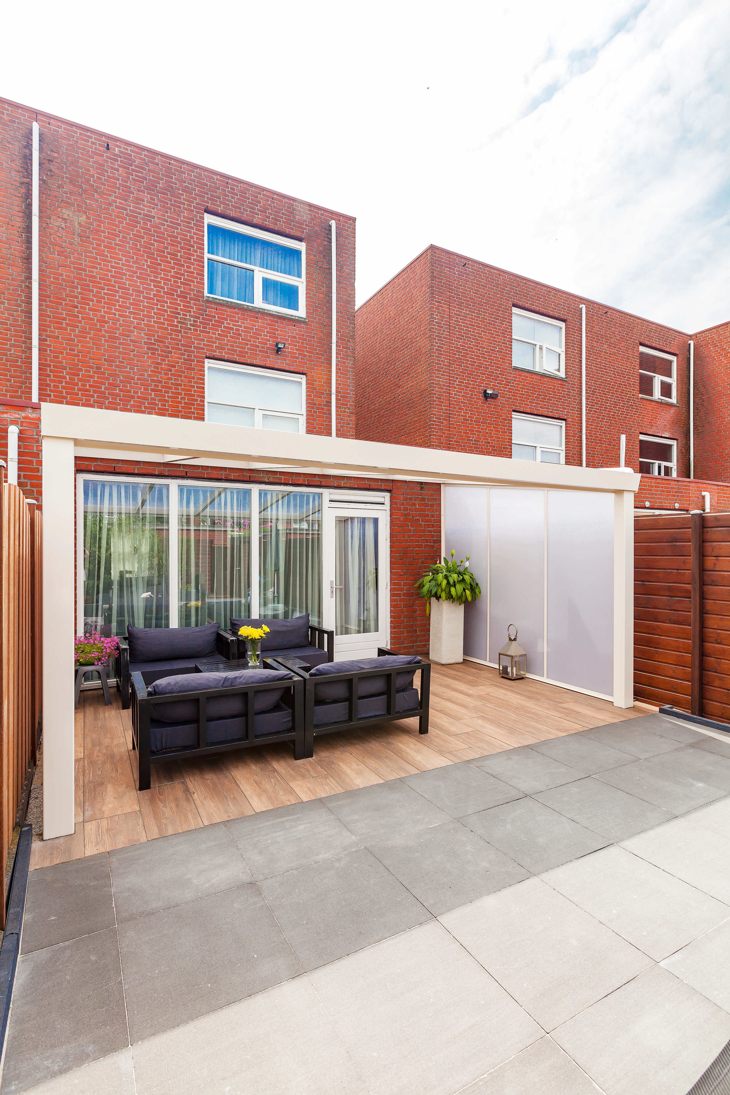 White-framed polycarbonate side wall enclosing a veranda corner with black sofa seating against a red-brick house